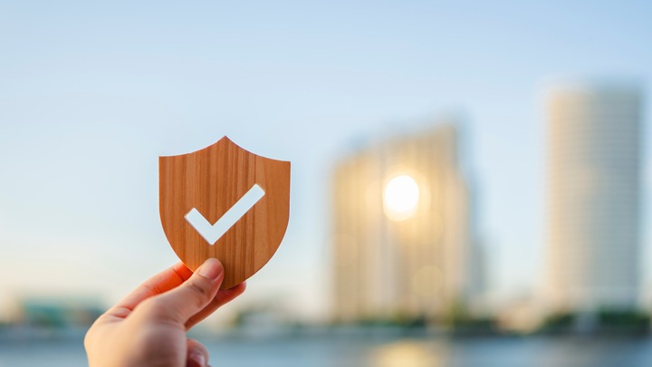 Bild zeigt Hand hält hölzernes Schild mit weißem Häkchen vor unscharfer Stadtkulisse verlinkt auf Blogbeitrag Risikolebensversicherung als Hinterbliebenenabsicherung